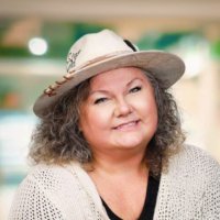 Debbie Sinclair smiling warmly, wearing a hat and cardigan in a soft, cozy portrait.